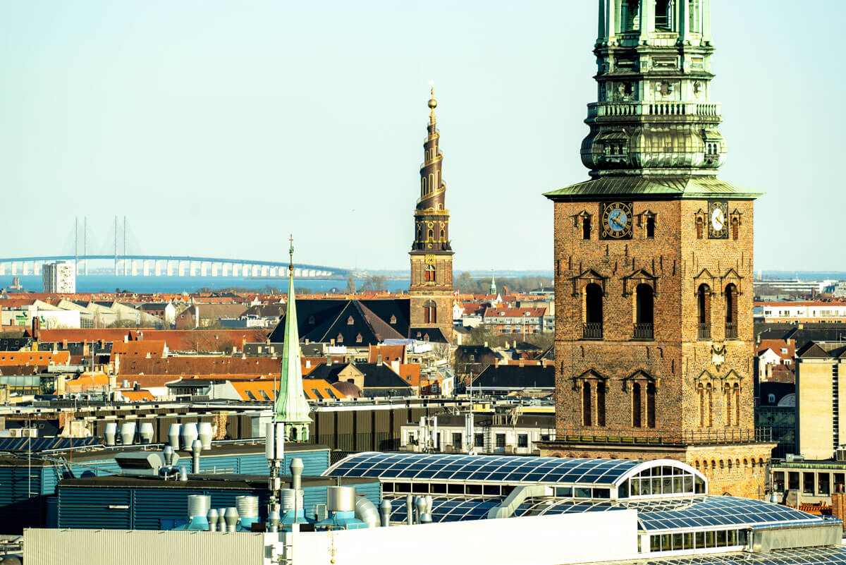 Vom Dach eines alten Turmes im Stadtzentrum von Kopenhagen kann man über die Dächer der Altstadt blicken. Am Horizont sieht man die Öresundbrücke mit den typischen 2 Pylonen