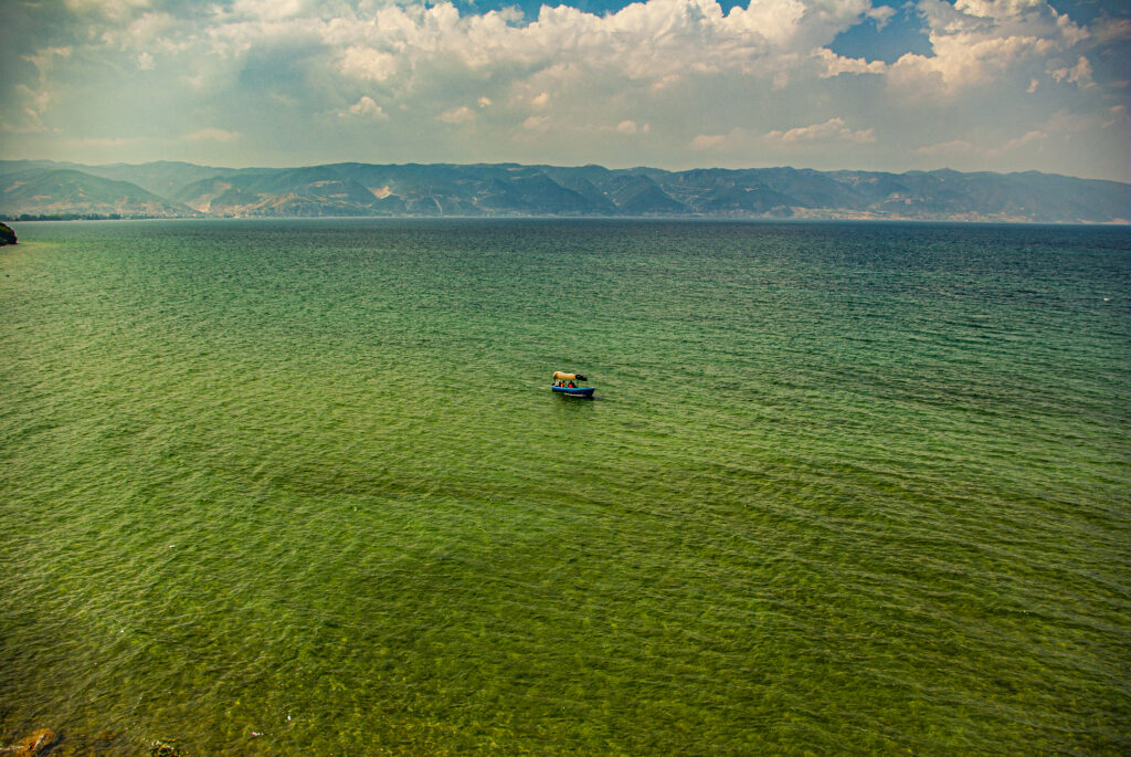 Blick über den Ohridsee. Auf dem See fährt ein kleines Boot. Im Hintergrund befinden sich hohe Berge.