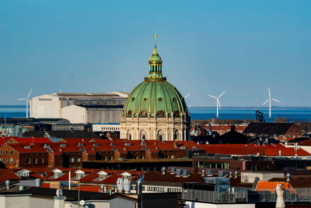 Die Kuppel der Frederiks Kirke ragt über die Dächer von Kopenhagen heraus. Im Hintergrund ist ein Windpark auf der Ostsee zu sehen.