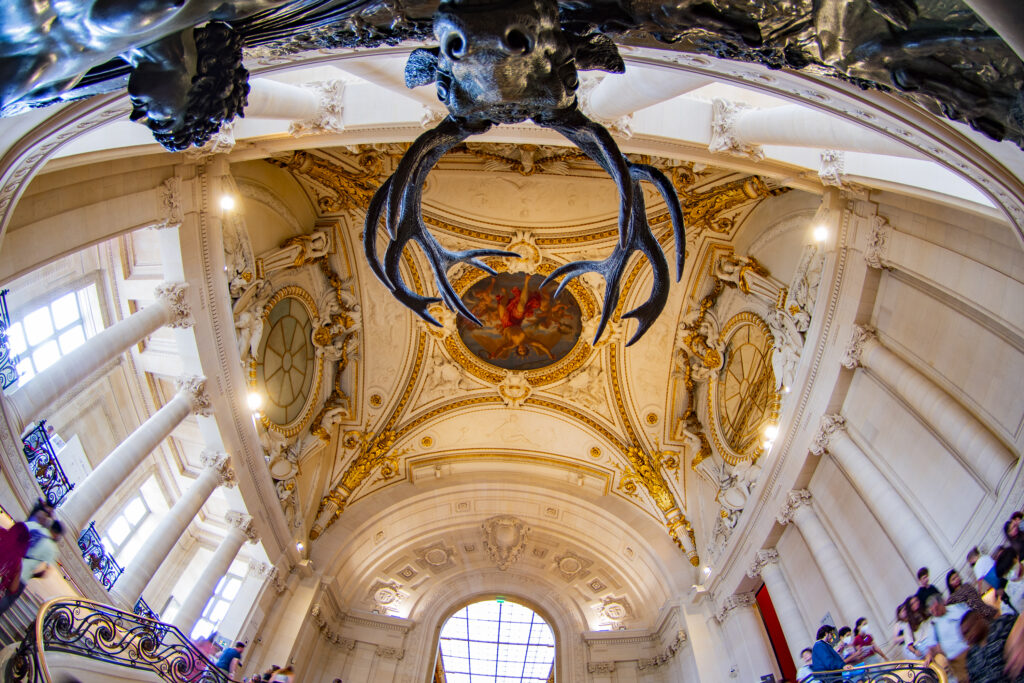 Galerie im Louvre, die die einzelnen Hallen verbindet. Der Blick ist auf die Decke der Halle gerichtet, die kunstvoll verziert ist. Riesige Mamorsäulen halten das Kuppelgewölbe.