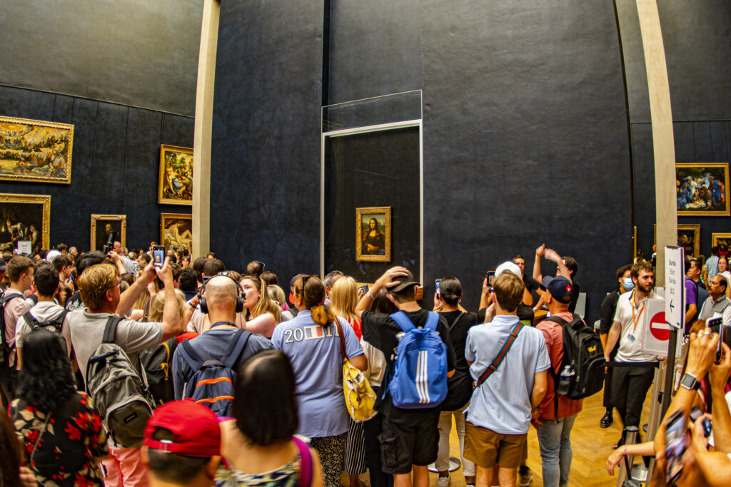 Blick auf die Mona Lisa im Louvre, die sich im Hintergrund auf einer schwarzen Wand präsentiert. Im Vordergrund sind zahlreich Menschen versammelt, die das Gemälde betrachten.