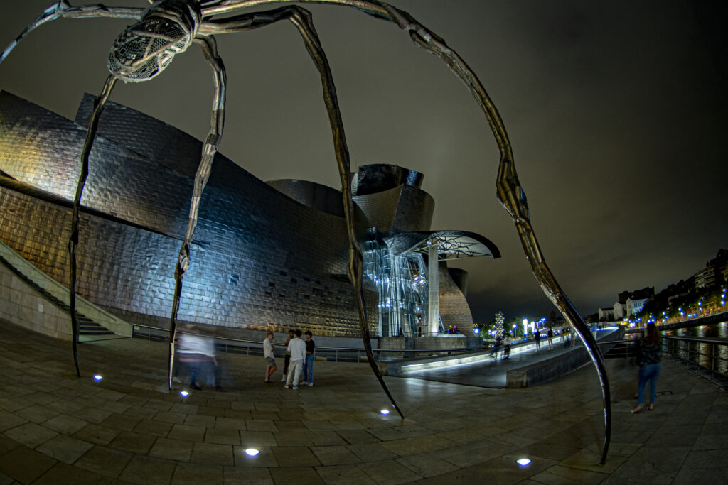 Die Nachtaufnahme des Guggenheimmuseums in Bilbao. Licht bricht an der Fassade des Gebäudes. Im Vordergrund ist die übergroße Plastik aus der Spinnen-Serie der Künstlerin Louise Bourgeois zu sehen.
