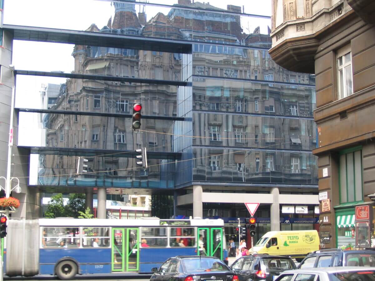 Straßenbild in Budapest mit enger Häusergasse. Im Bild ein moderne Glasbau in dessen Fassade sich ein altes Gebäude spiegelt.