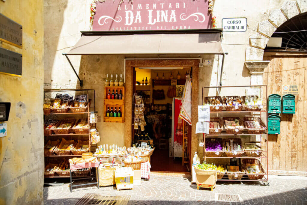 Der Eingang eines Delikatessenladens in der Alstadt von Malcesine. Reschts und links vom Eingang sind Regale und Körbe aufgestellt gefüllt mit Tüten und Flaschen.