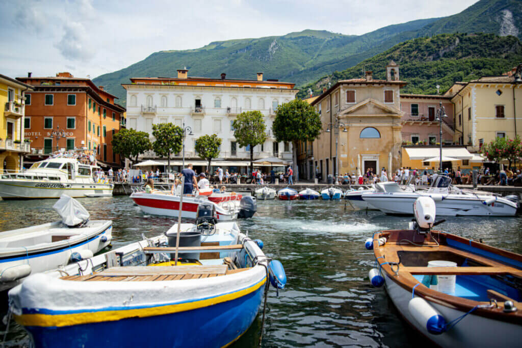 Im Hafenbecken vom Malcesine liegen kleine Boote. Im Hintergrund befindet sich die Promenade von Bäumen und alten mehrstöckigen Gebäuden gesäumt. Vor den Häusern laufen viele Menschen auf der Straße oder sitzen in Cafes.