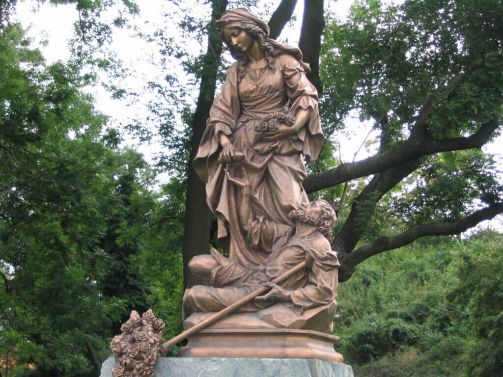 Die lebensgroße Skulptur von Elisabeth von Thüringen steht auf einem Granitsockel im Park zwischen Bäumen.