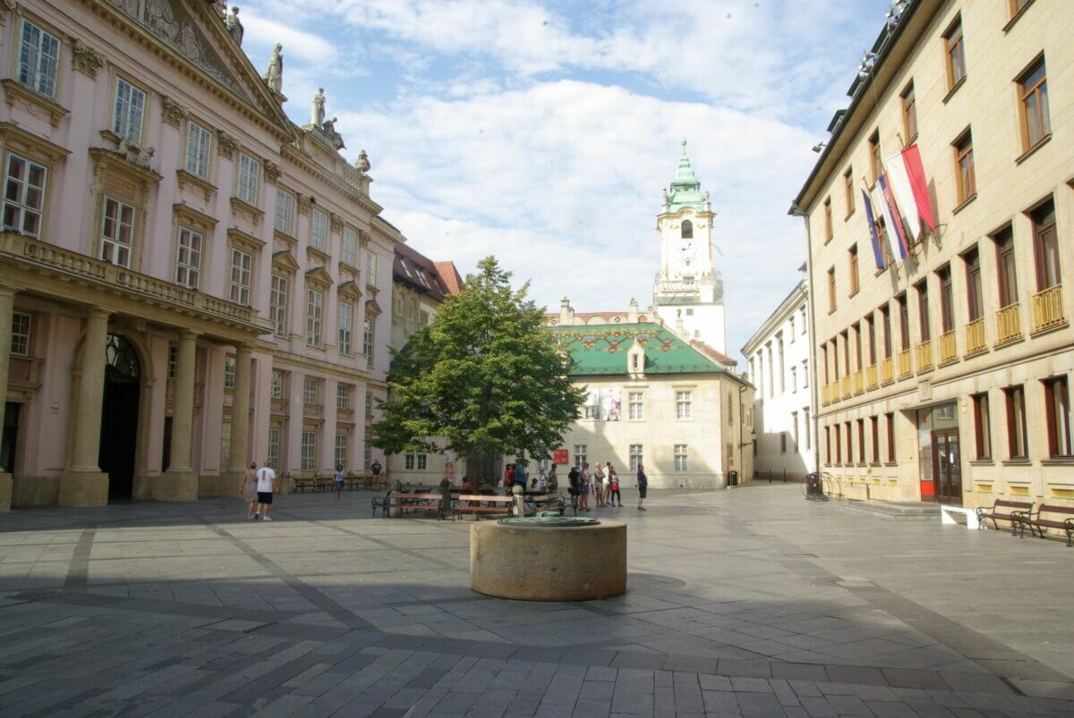 Ein gepflasterter Platz mit alten Gebäuden in Bratislava. Im Hintergrund ist der Turm des Alten Rathauses mit einer grünen Spitze zu sehen.