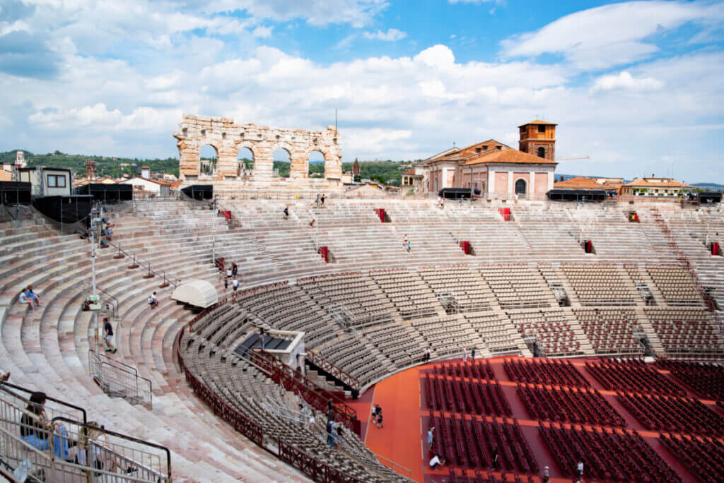 Das alte Amphitheater in Verona von den oberen Plätzen aus fotografiert. Im Innenraum sind die Stühle für die opernbesucher zu sehen und am oberen Rand der Arena kann man die Dächer der Altstadt erkennen. Der Himmel ist blau.