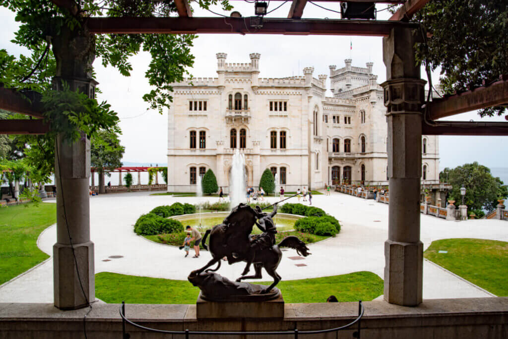 Aus einer Pergola das Parks heraus fotografiert sieht man das Schloss. Davor in der Mitte ein Springbrunnen und eine Skulptur von einem Reiter auf einem Pferd.