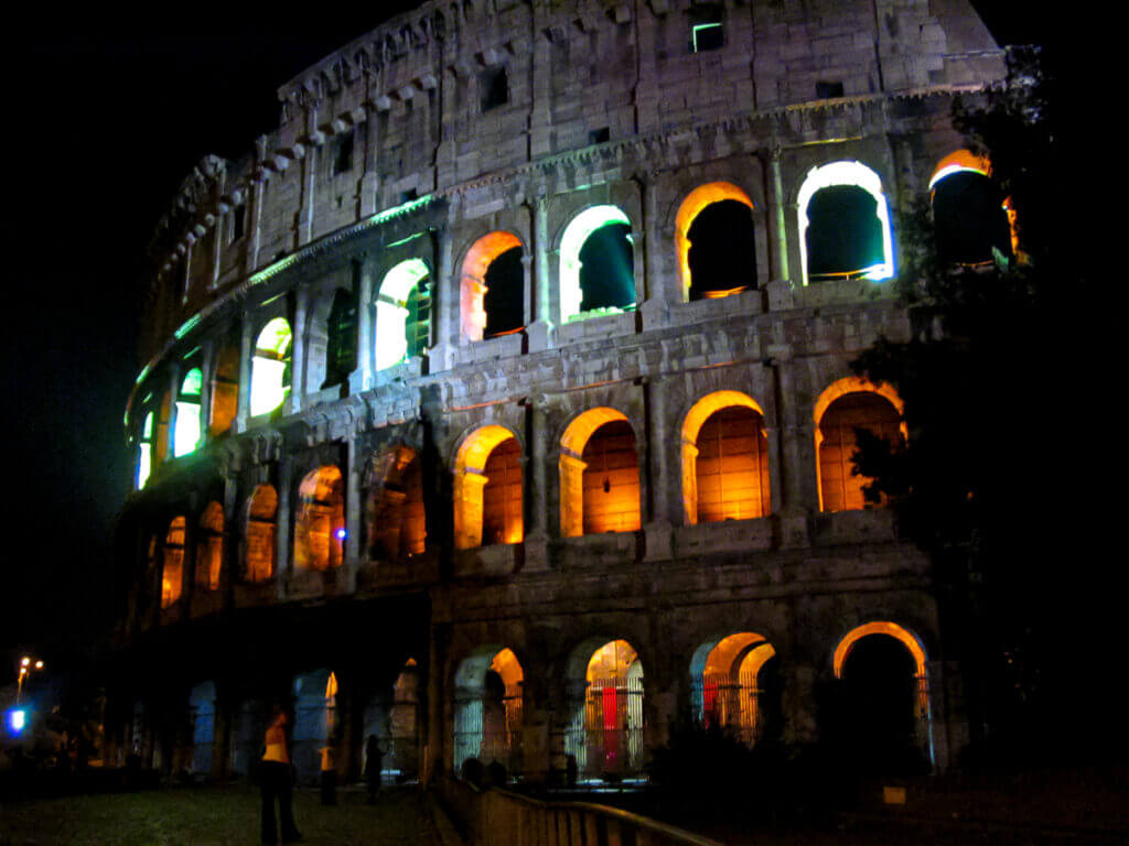 Die Fassadenruine des Kolosseum bei Nacht. Drei Etagen hoch mit aneinander gereihten Rundbögen, die von innen erleuchtet sind.