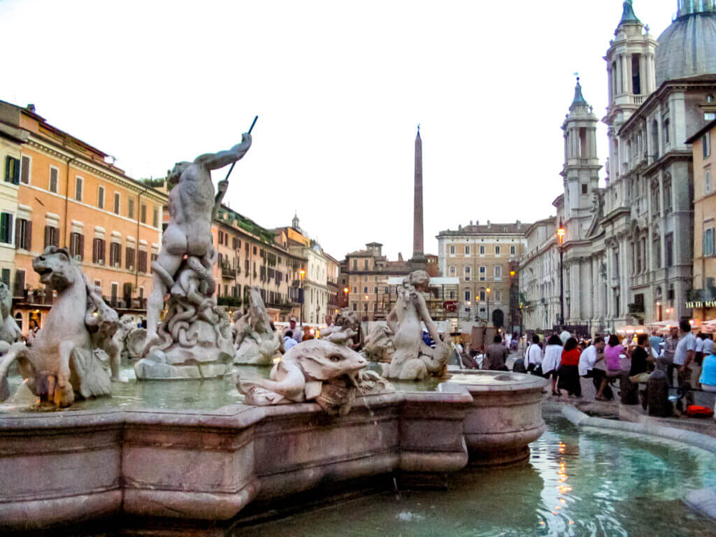Im Vordergrund ist ein Schmuckbrunnen in Rom zu sehen in dessen Mitte eine nackte männlich Figur mit einem Dreizack steht. Weitere Skulpturen umrahmen den Brunnen. Im Hintergrund sieht man einen Platz von Hausfassaden eingegrenzt.