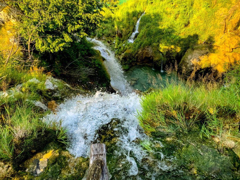 Ein mit grünen Gräsern umsäumter Wasserfall. Mit Blick von ober nach unten ergibt sich der Eindruck, dass das Wasser in der Bildmitte verschwindet.