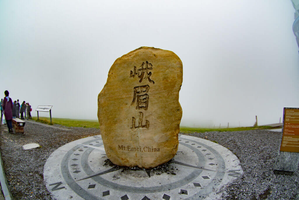 Zu sehen ist ein Basaltstein aus China der hellgelb bis braun schimmert. Darauf sind drei chinesische Schriftzeichen zu sehen mit einem Schriftzusatz "Mt.Emei, China". Der Stein steht auf einer Windrose aus Stein.
