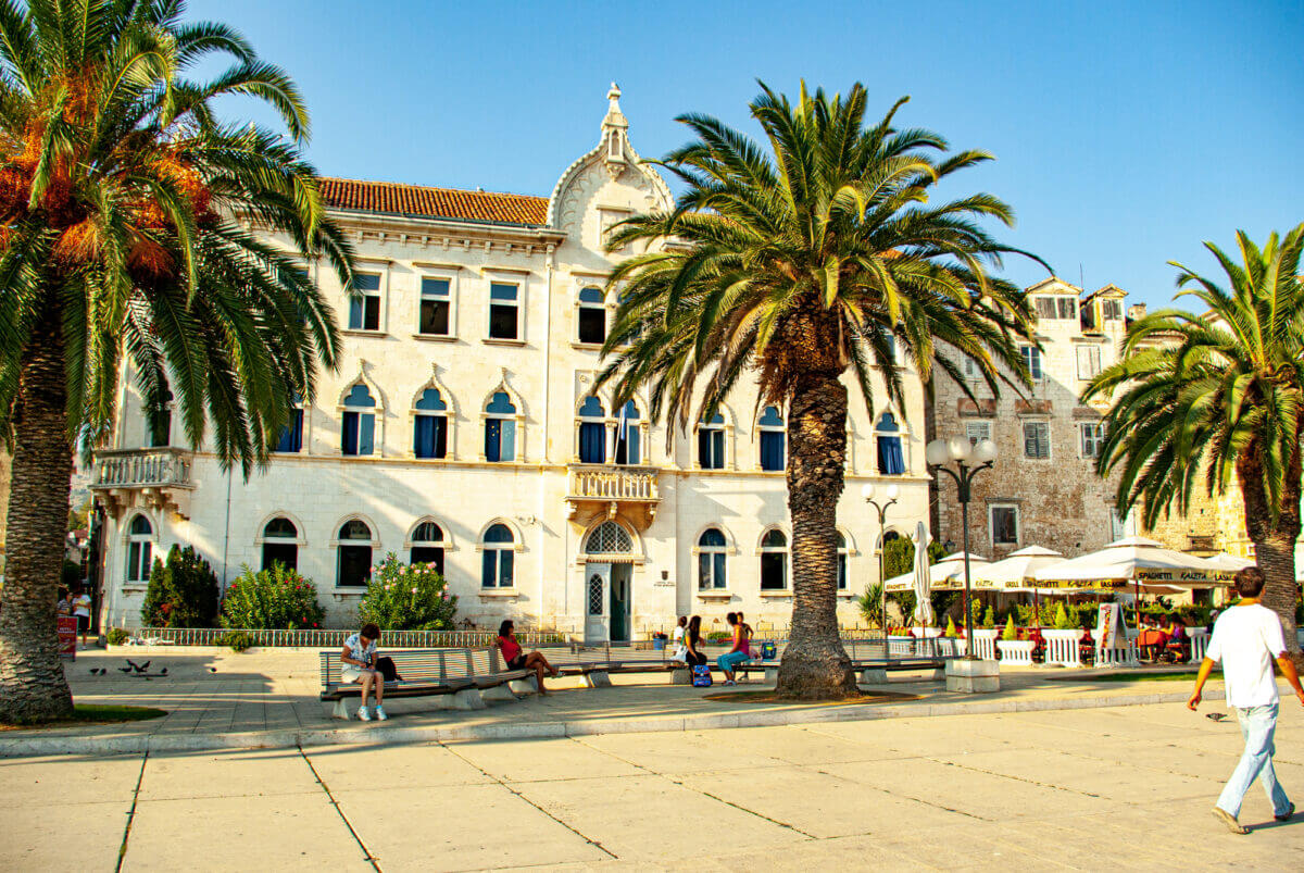 Die Fassade der Petar-Berislavić-Grundschule in Trogir ist auf dem Bild zu sehen. Davor stehen Palmen und Bänke auf denen menschen sitzen.