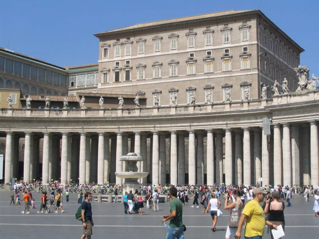 Zu sehen ist die Cappella Niccolina ein mehrstöckiges Haus direkt am Petersplatz. Im Vordergrund ist der Säulengang des Petersdoms zu sehen und die Menschenschlangen, die auf den Einlass in den Dom warten.