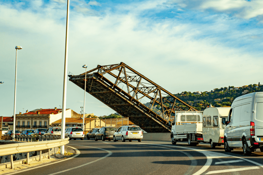 Die Pont Maréchal-Foch in Sète ist eine Eisenbahnklappbrücke aus Stahl, welche im Moment der Aufnahme hochgefahren ist. Im Vordergrund stehen zahlreiche Autos auf der Straße, die an der Brücke vorbei führt.