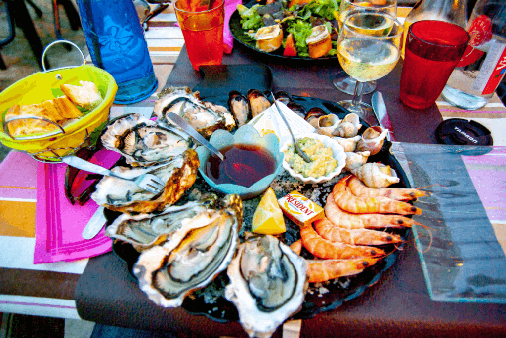 Auf dem Foto ist ein großer Teller mit Auster, Garnelen und Muscheln in einem Restaurant in Sète zu sehen. Dazu stehen Brotkorb, Wein- und Wasserglas.