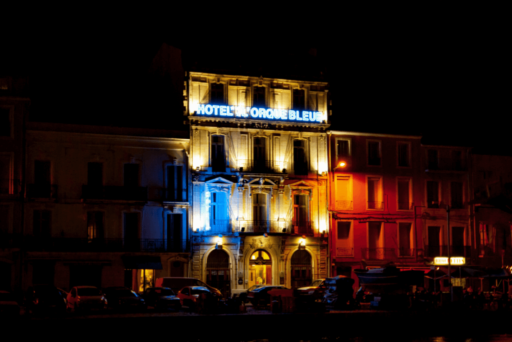 Eine Nachtaufnahme von der Hotelfassade des Hôtel l'Orque Bleue in Sète. Sie ist in den Farben blau, weiß und rot angeleuchtet.