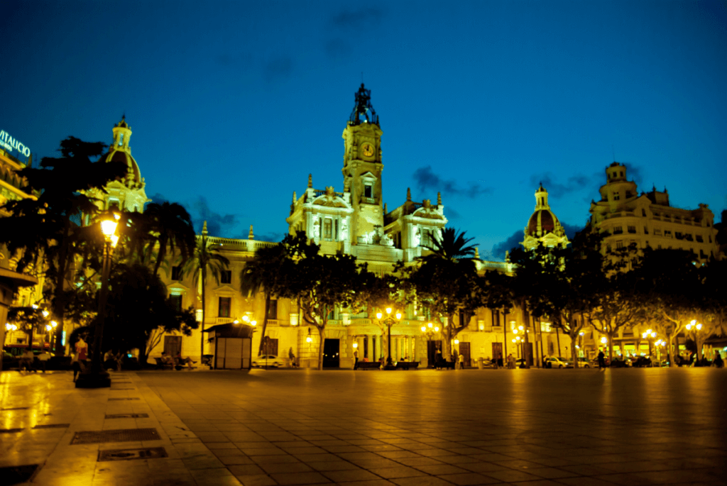 Das Foto zeigt die Aussenansicht des Rathauses von Valencia kurz vor Einbruch der Nacht. Vor dem Gebäude stehen Bäume und davor ist ein großer offener Platz.