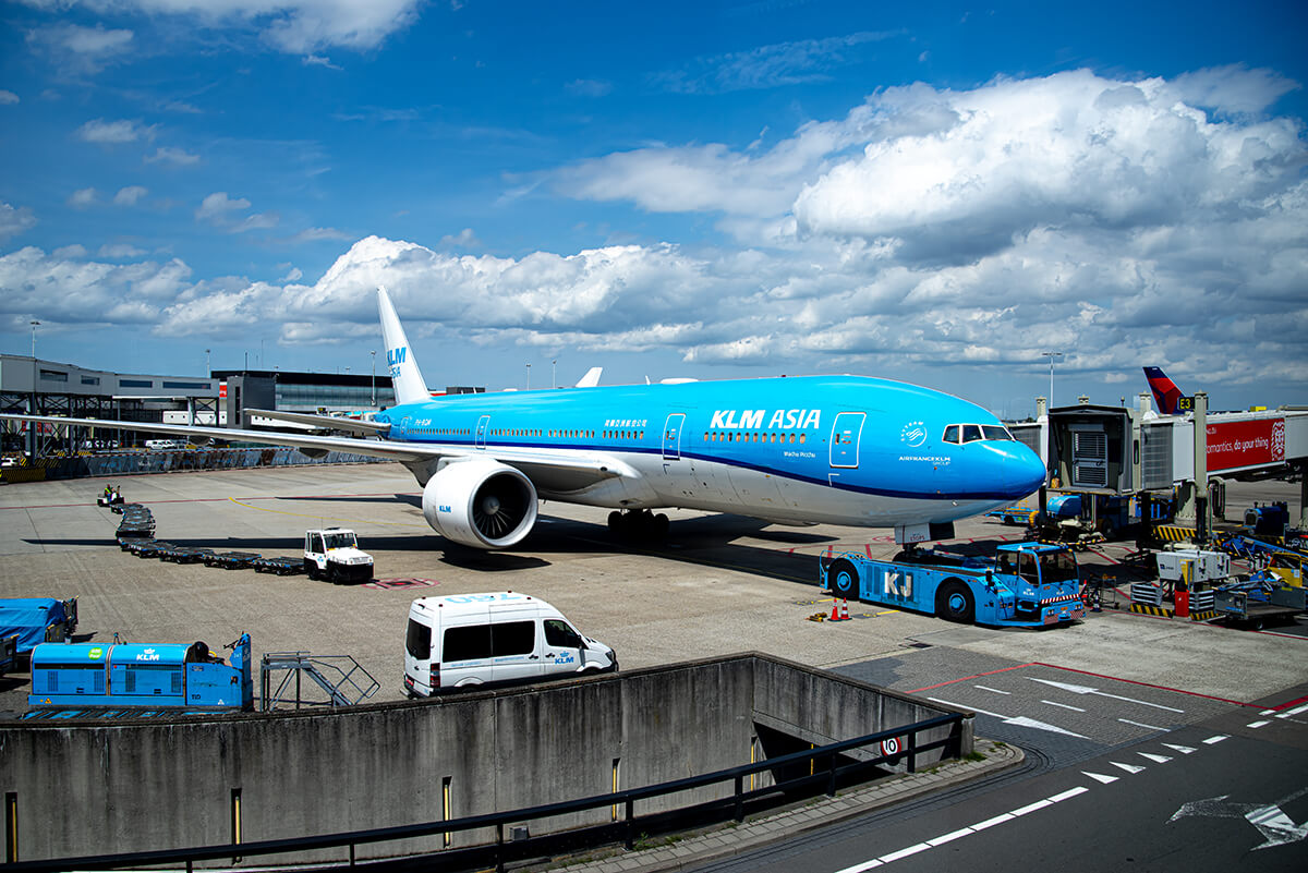 Reisezeit für ein blau-weißes Flugzeug? Es steht am Gate eines Flughafens. Drum herum stehen Gepäckwagen und Autos.