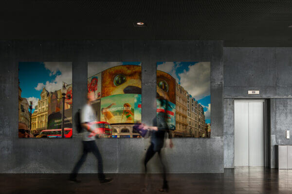 Der Piccadilly Circus ist berühmt für seine gebogene haushohe Leuchtreklametafel, die hier im Mittelpung des Bildes zu sehen ist. Von links durchquert ein typischer roter Doppelstockbus das Bild. Zu sehen in einem Bilderrahmen