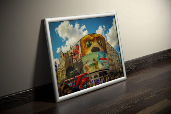 Der Piccadilly Circus ist berühmt für seine gebogene haushohe Leuchtreklametafel, die hier im Mittelpung des Bildes zu sehen ist. Von links durchquert ein typischer roter Doppelstockbus das Bild. Zu sehen in einem Bilderrahmen