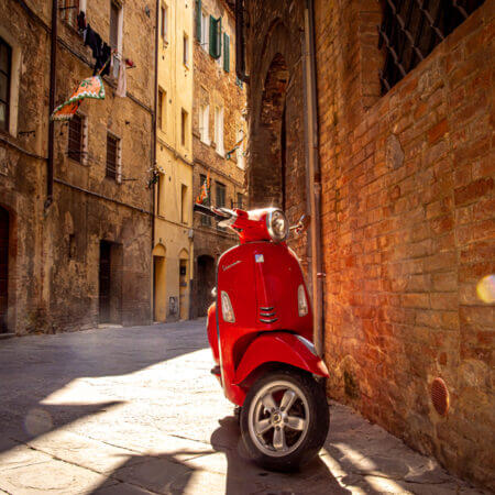 In den Gassen Sienas zwischen gemauerten Fassaden steht einsam eine rote Vespa. Die Sonne blitzt auf dem verchromten Lenker auf.