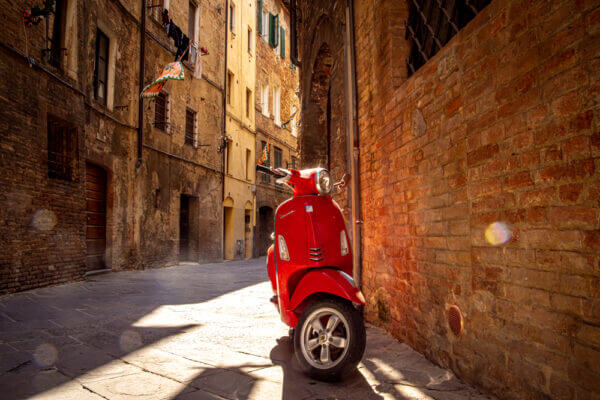 In den Gassen Sienas zwischen gemauerten Fassaden steht einsam eine rote Vespa. Die Sonne blitzt auf dem verchromten Lenker auf.