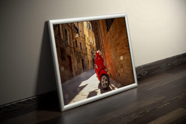 In den Gassen Sienas zwischen gemauerten Fassaden steht einsam eine rote Vespa. Das Bild ist in einem Bilderrahmen eingerahmt.