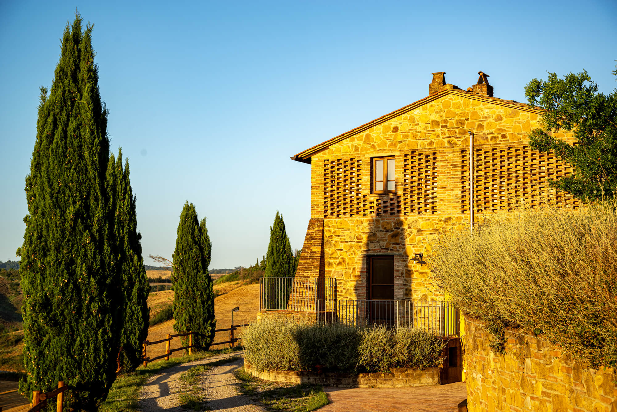 Ein Haus aus Naturstein, eingerahmt in Zypressen und einer Hügellandschaft in der Toskana. Im morgendlichem Licht.