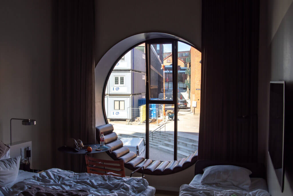 Ein Hotelzimmer mit Blick durch ein rundes Fenster. Die Betten sind unordentlich und der Blick richtet sich auf ein Kontenerhaus.