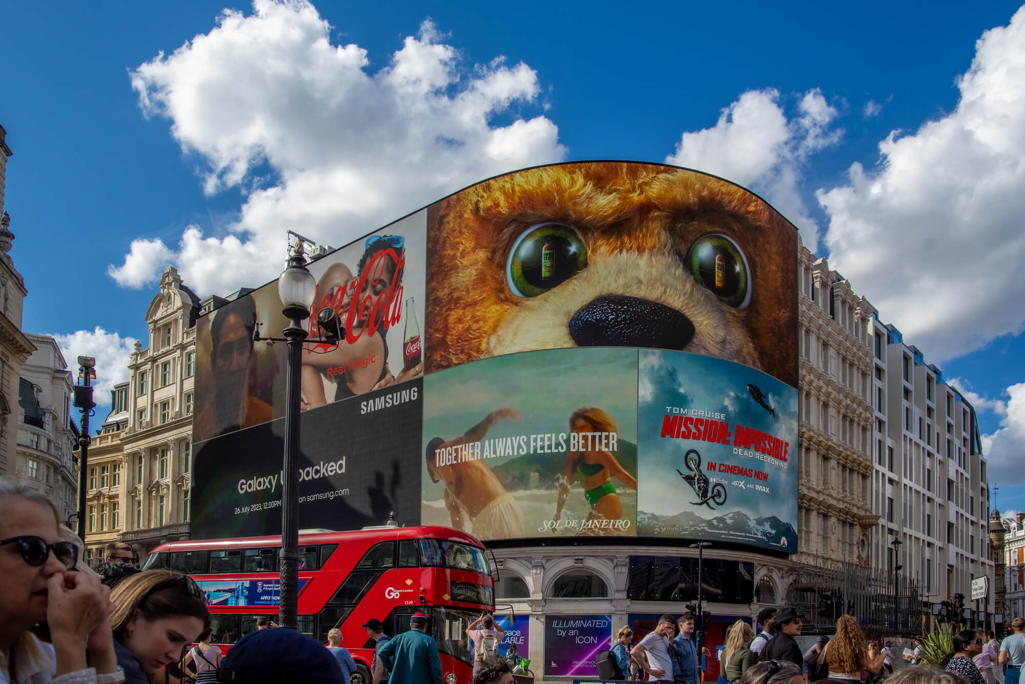 Der Piccadilly Circus ist berühmt für seine gebogene haushohe Leuchtreklametafel, die hier im Mittelpung des Bildes zu sehen ist. Von links durchquert ein typischer roter Doppelstockbus das Bild.