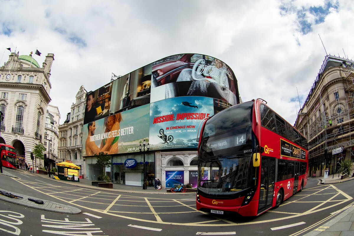 Der Piccadilly Circus ist berühmt für seine gebogene haushohe Leuchtreklametafel, die hier im Mittelpung des Bildes zu sehen ist. Von rechts durchquert ein typischer roter Doppelstockbus das Bild.