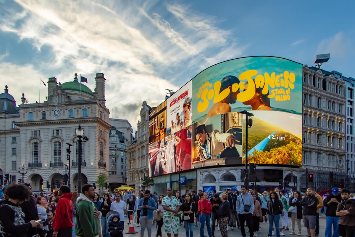 Der Piccadilly Circus ist berühmt für seine gebogene haushohe Leuchtreklametafel, die hier im Mittelpung des Bildes zu sehen ist. Im Vordergrund stehen viele Menschen.