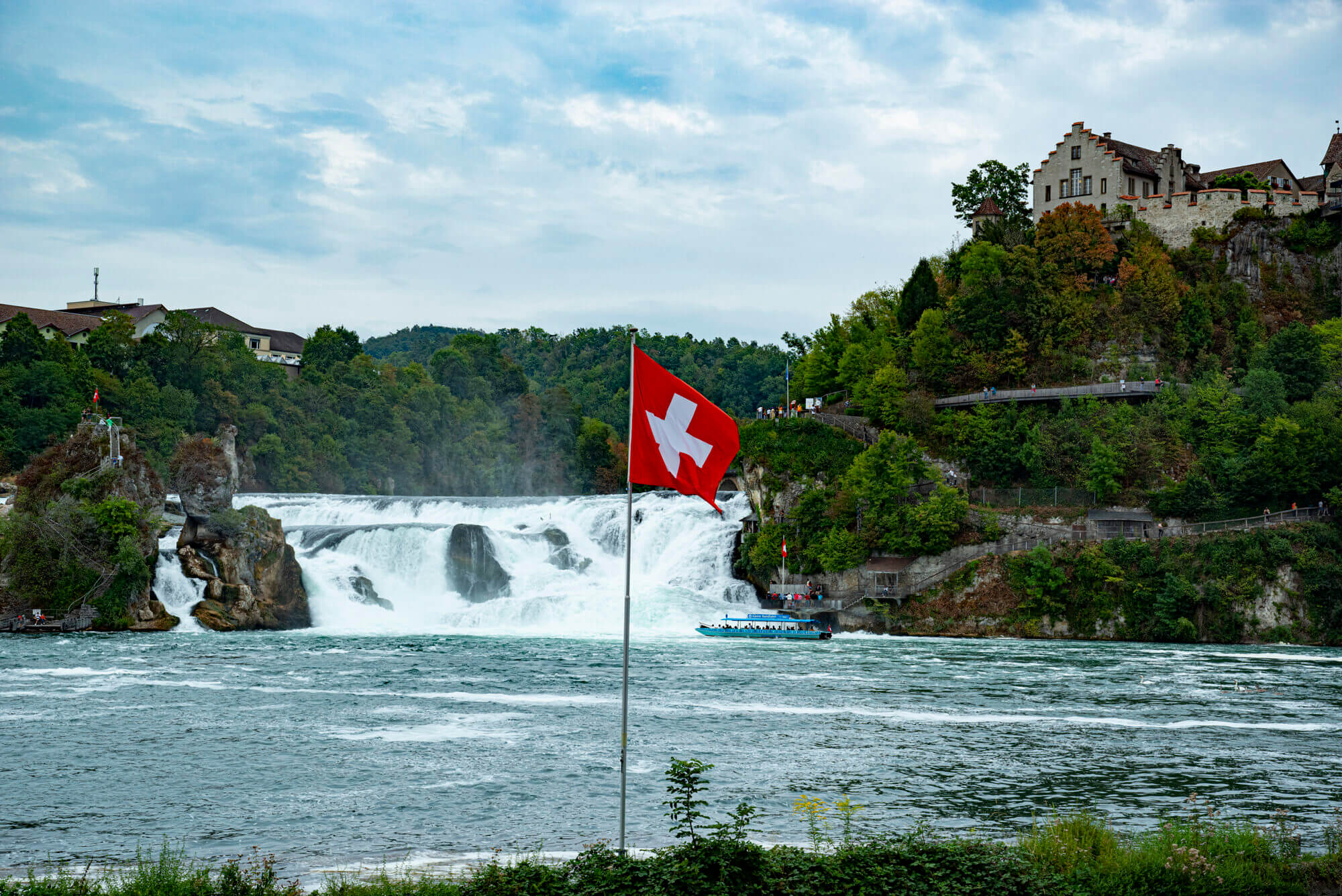 Vom Westufer des Rheins in Richtung des Schloß Laufen blickend schaut man den Wasserfall hinauf. Im Vordergrund steht ein Fahnenmast mit Schweizer Flagge.