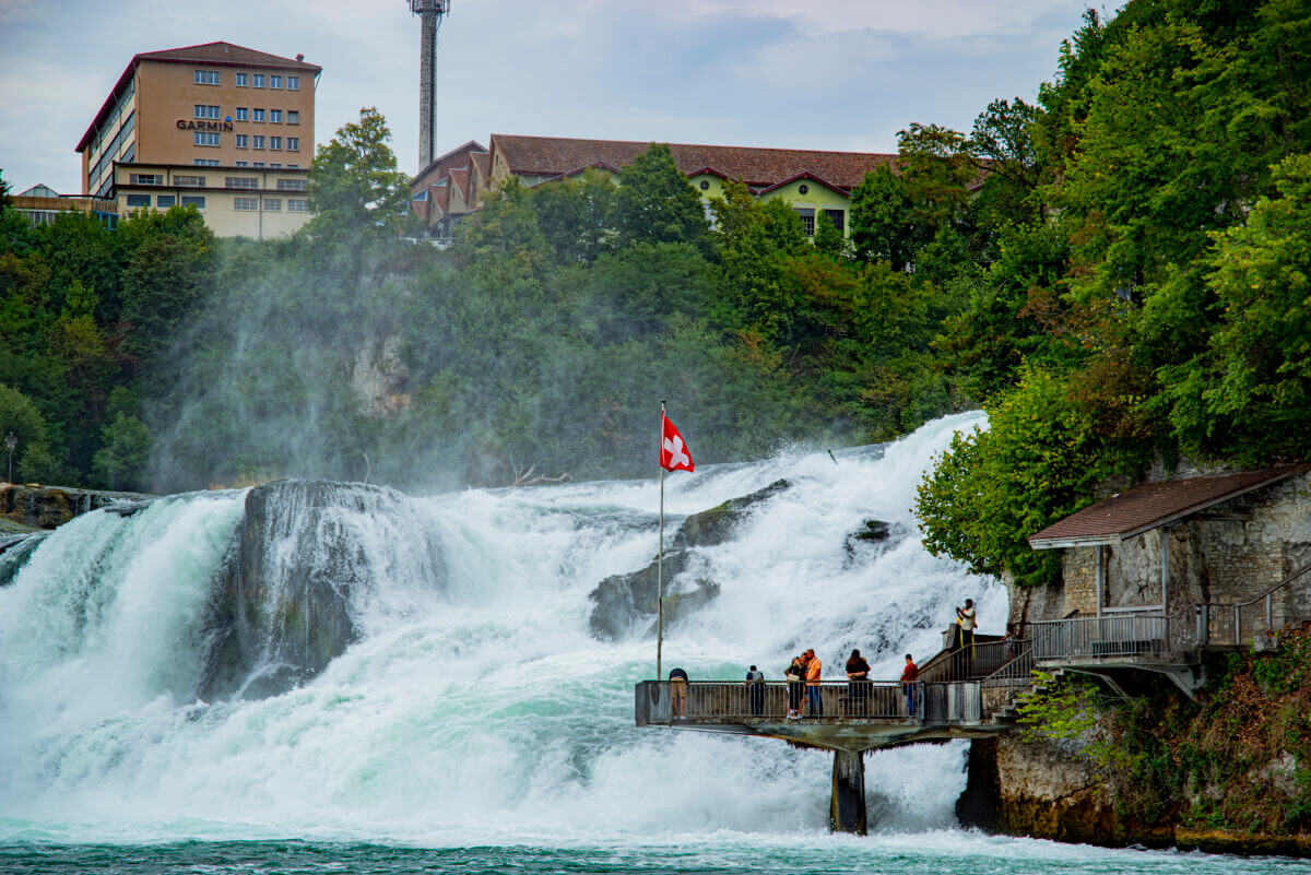 Vom Westufer des Rheins in Richtung der Wasserfälle blickend sieht man davor die Besucherplattform - eine Art Steg der dierekt vor die Wasserfälle führt. Darauf befindet sich ein Fahnenmast mit Schweizer Flagge.