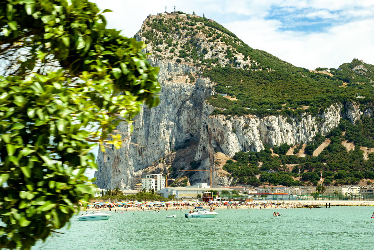 Foto vom Felsen Gibraltars. Davor ist die Bucht und der Stand zu sehen mit zahlreichen Badegästen. Es ist Sommer.