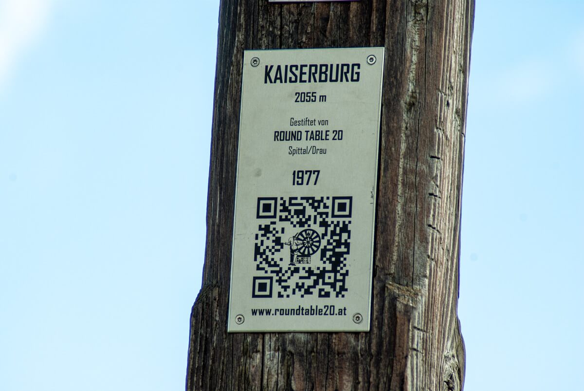 An einem Holzmast befestigte Hinweistafel zur Kaiserburg. Mit der Information, dass der Gipfel 2055 Meter hoch ist.