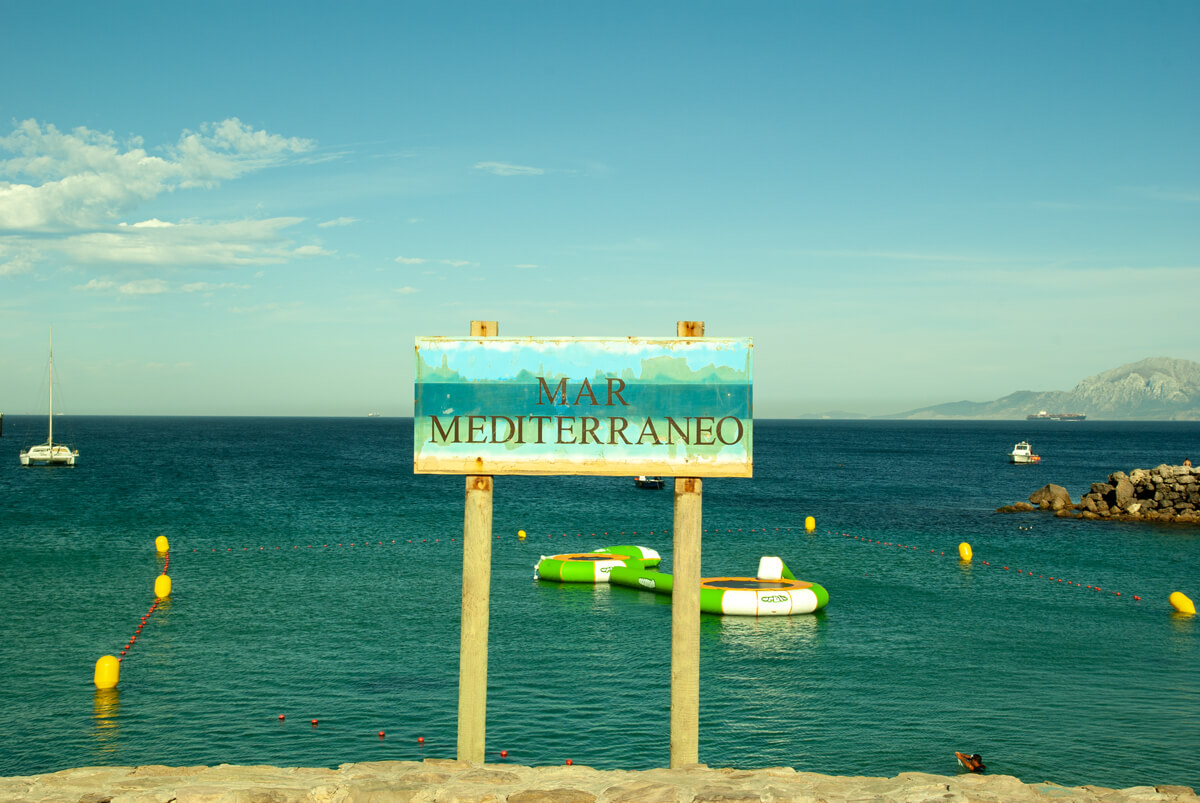 Auf dem Foto ist ein rechteckiges Holzschild auf zwei Holzstielen mit der Aufschrift MER MEDITERRANEO zu sehen. Im Hintergrund sieht man das Mittelmeer und einzelne kleine Boote.