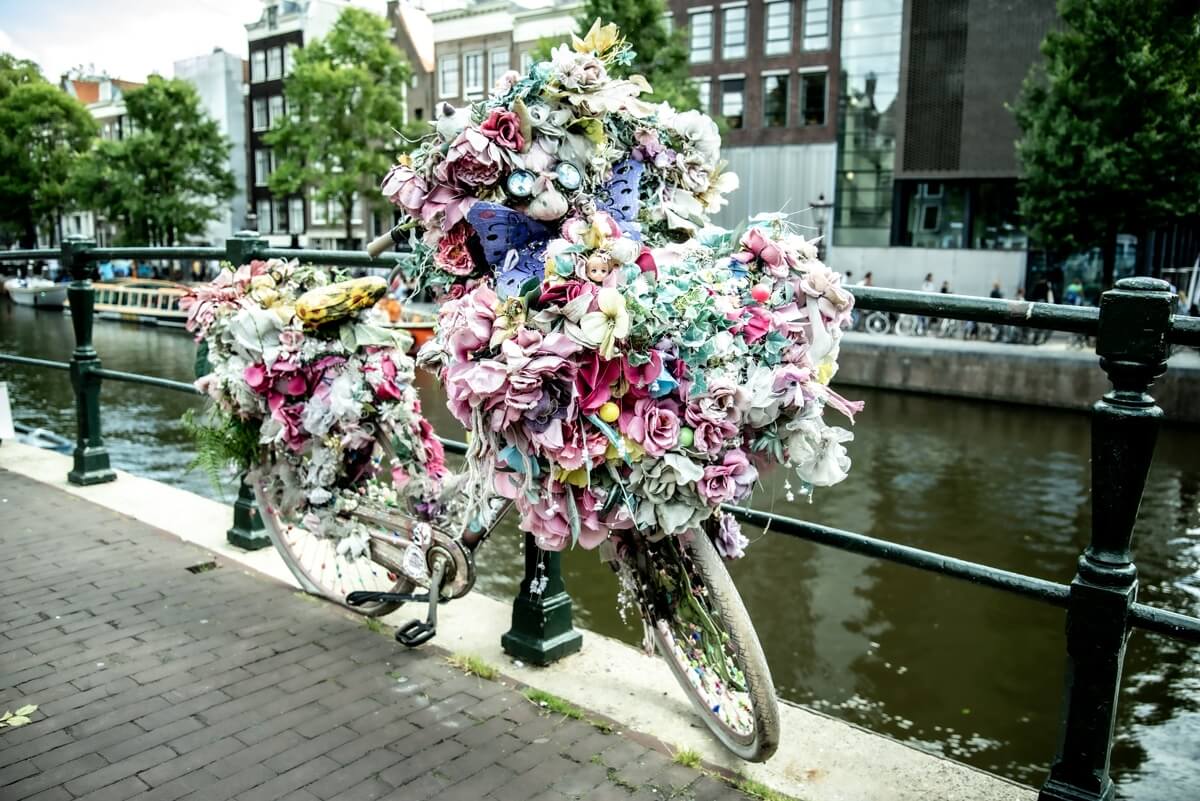 Ein mit Kunstblumen dekoriertes Fahrrad in Amsterdam, welches an einem Geländer an einen der zahlreichen Wassergräben angeschlossen ist.