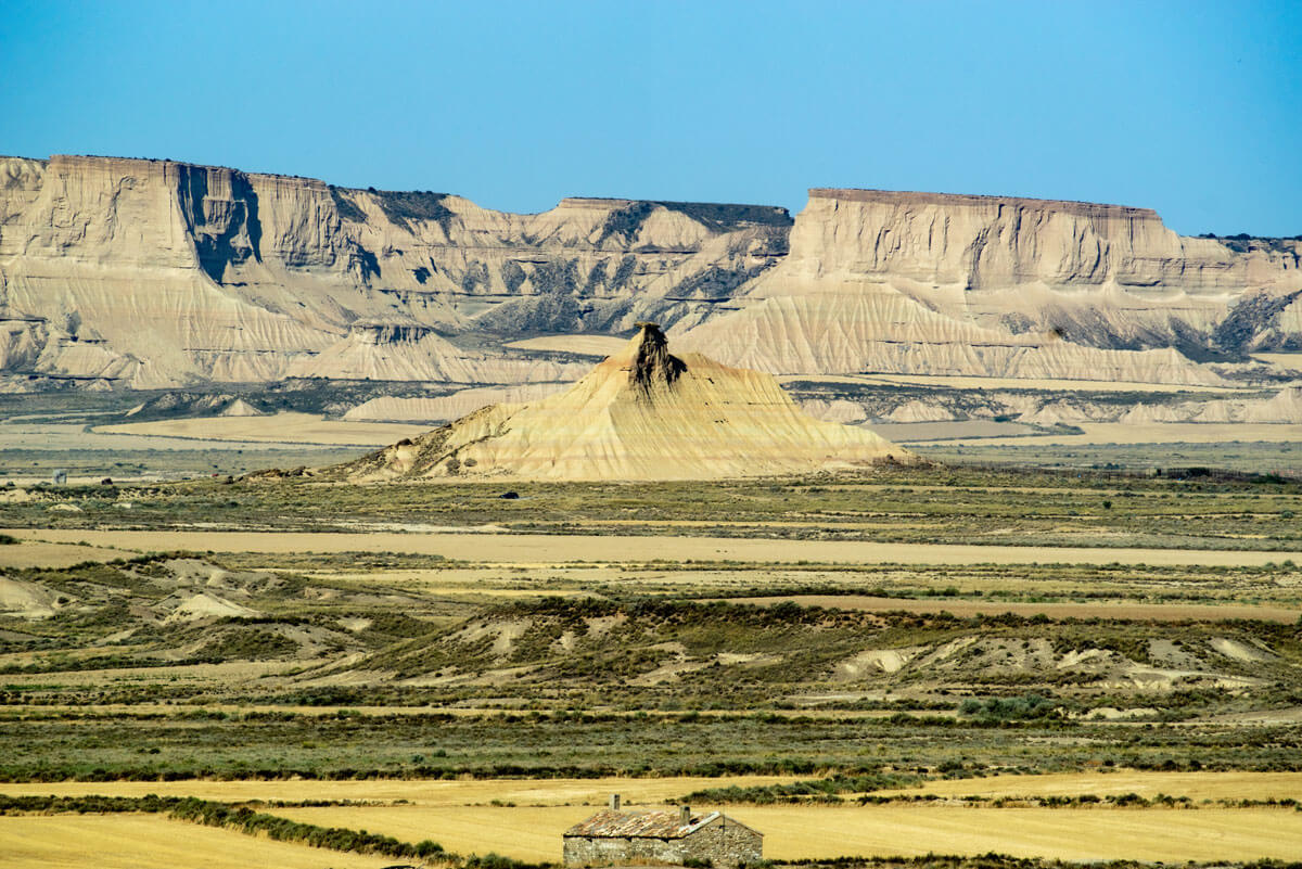 Von einer Anhöhe in der Halbwüste aus erblickt man die weite zerklüftete Landschaft. Sandsteinformationen und trockene Pflanzen. In der Bildmitte steht e