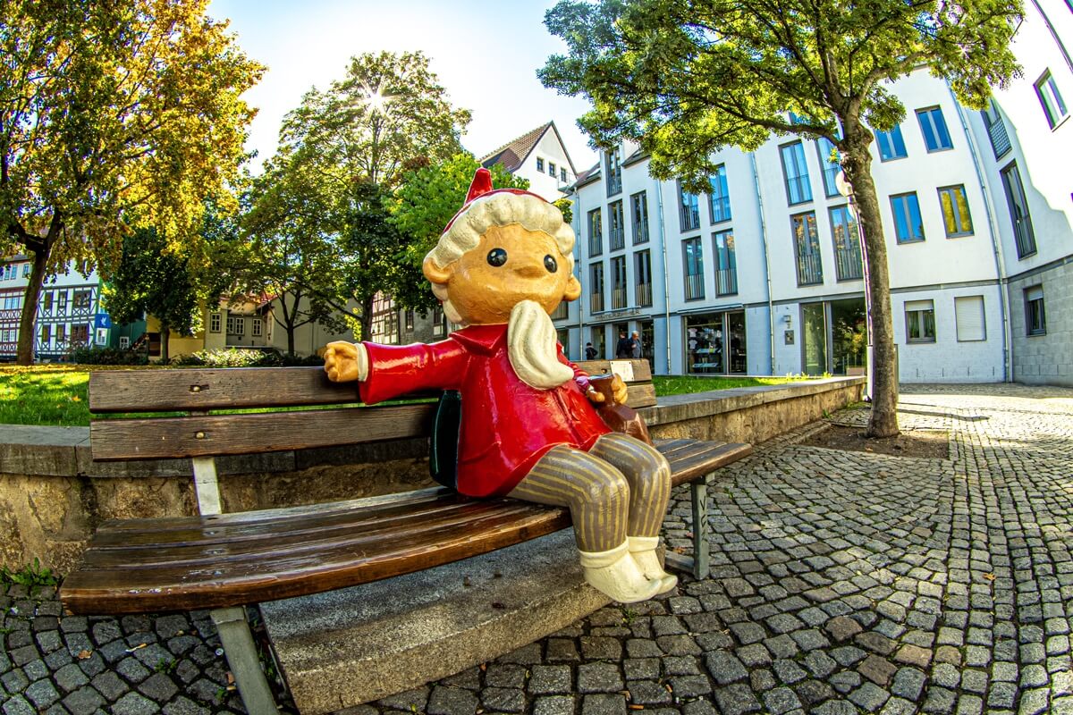Erfurt ist eine Fernsehstadt. Auf dem Bild ist das Sandmännchen in lebensfgröße sitzend auf einer Parkbank zu sehen. Im Hintergrund stehen Bäume und die Sonne scheint durch die Blätter.