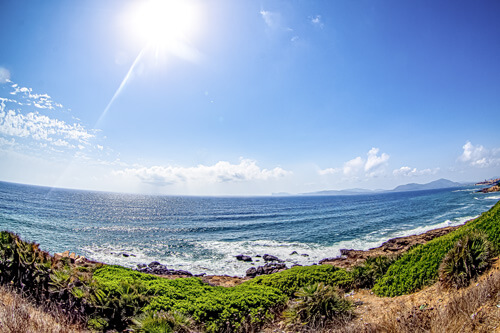 Zu sehen ist ein Strand auf Sardinien. Die Sonnsteht hoch am Himmel und die Wellen branden zwischen den Steinen am Strand.
